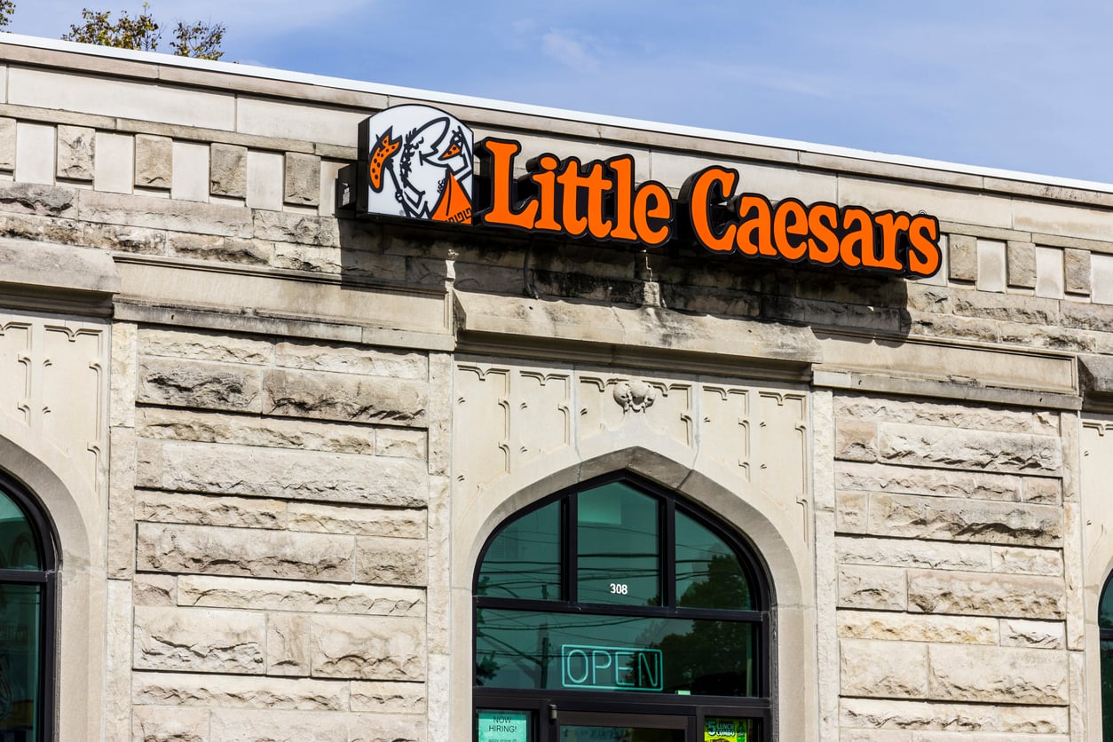 The exterior of a stone building with a “Little Caesars” sign above the entrance and an illuminated “OPEN” sign in the window below.