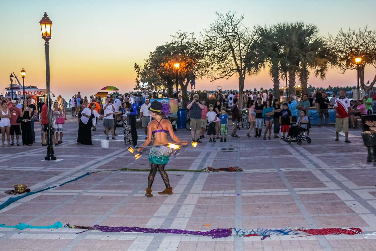 A street performer with hula hoops and fire entertains a crowd at sunset in a plaza lined with palm trees and lampposts. People watch, some taking photos, as colorful scarves lie on the ground.