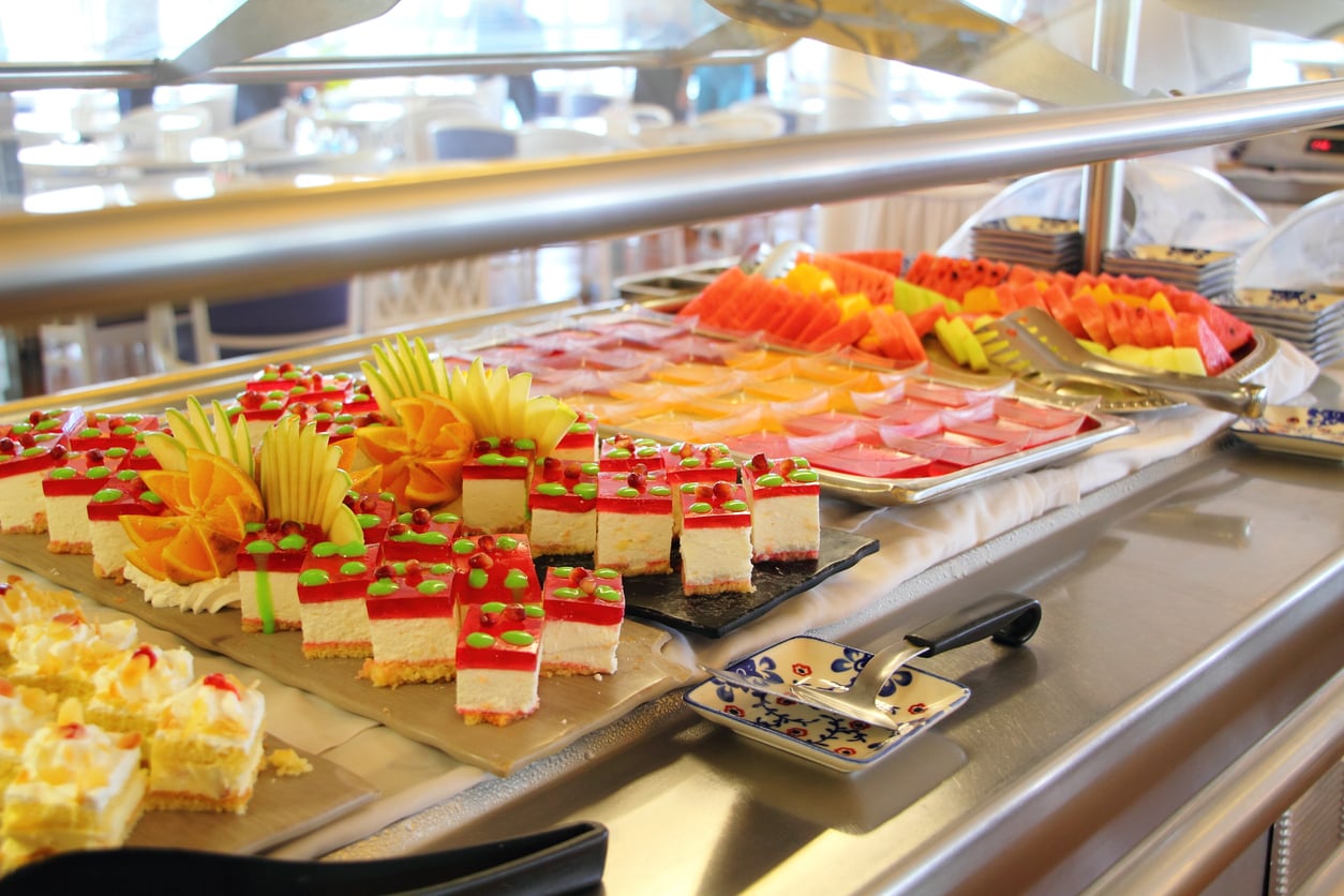 A buffet table with assorted desserts including colorful cakes topped with fruit, sliced watermelon, and other assorted fruit, all arranged neatly on trays with serving utensils.
