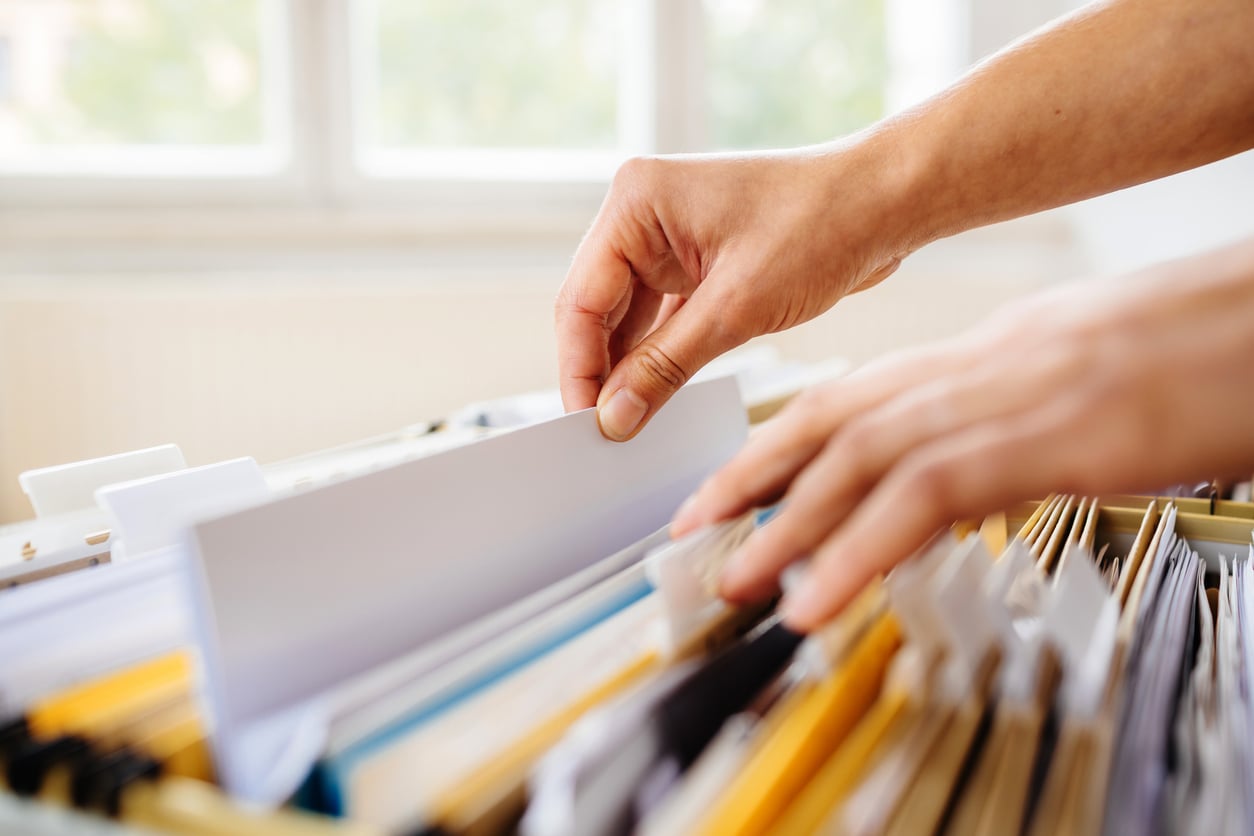 A person’s hands searching through a filing cabinet filled with folders and documents, in a well-lit room with windows in the background.