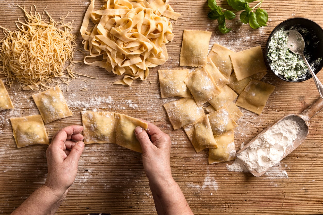 Hands making homemade ravioli on a wooden table, surrounded by uncooked pasta, flour, fresh basil, and a bowl of ricotta spinach filling.
