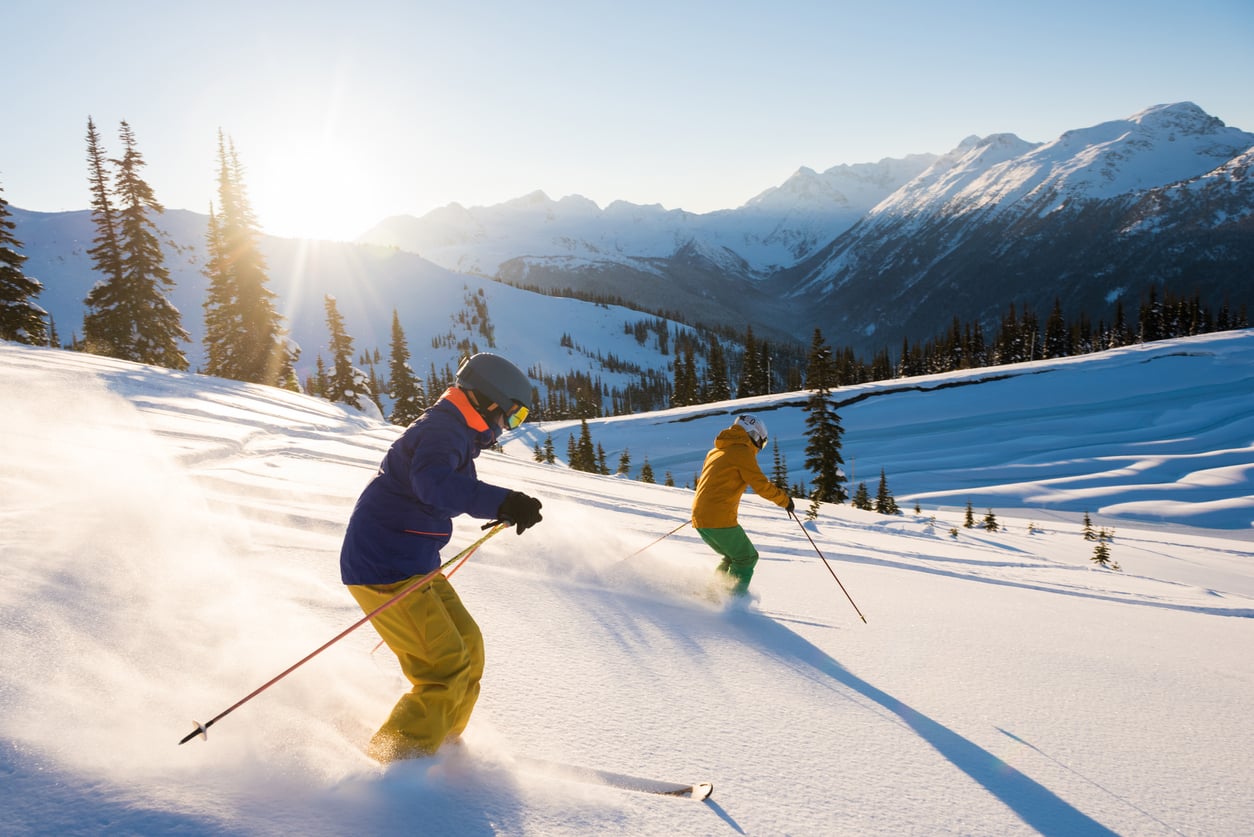 Two people skiing down a snowy mountain slope at sunrise, surrounded by snow-covered trees and distant mountain peaks under a clear sky.