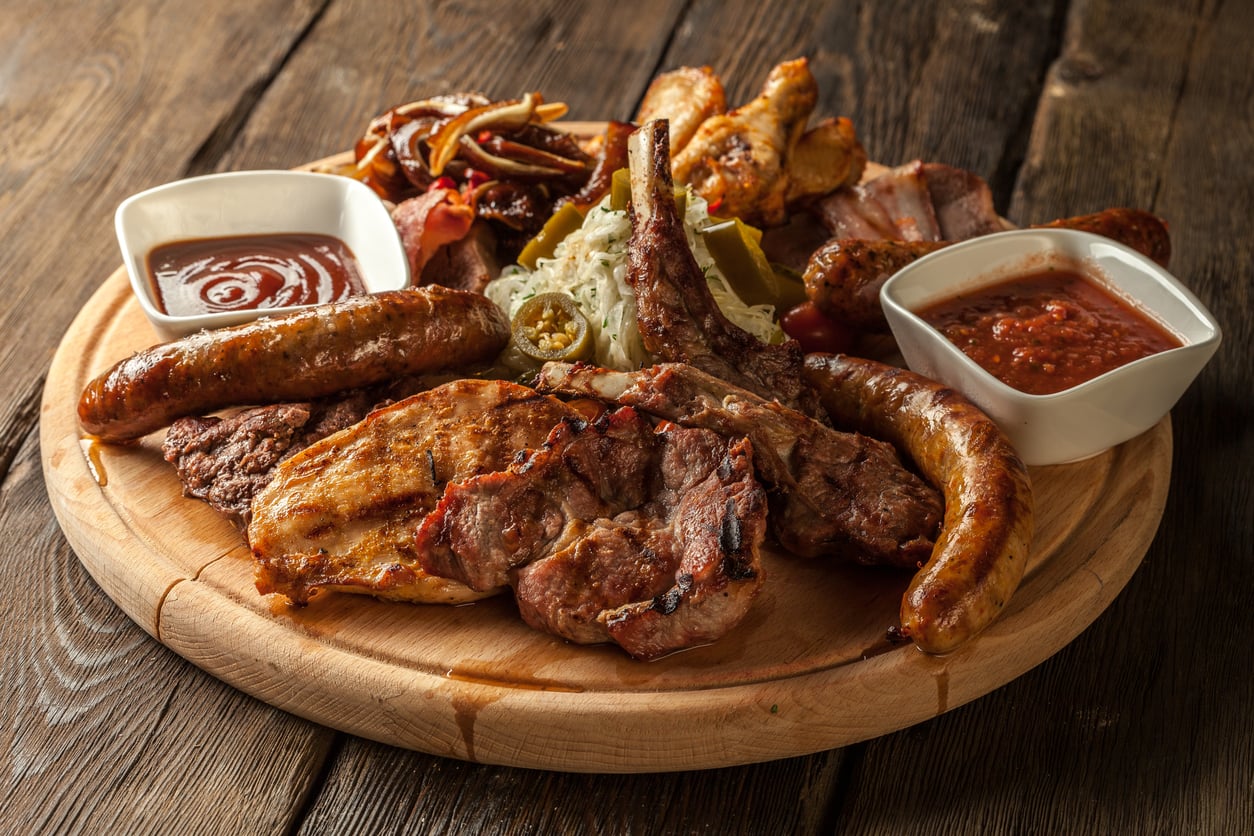 A wooden platter with grilled meats including sausages, ribs, and chicken, served with two sauces and a side of vegetables on a rustic wooden table.
