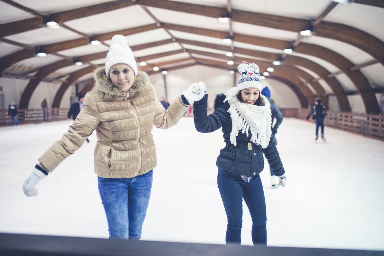 Two women in winter clothes ice skate together indoors, holding hands and smiling. Other skaters are visible in the background under the arched roof.