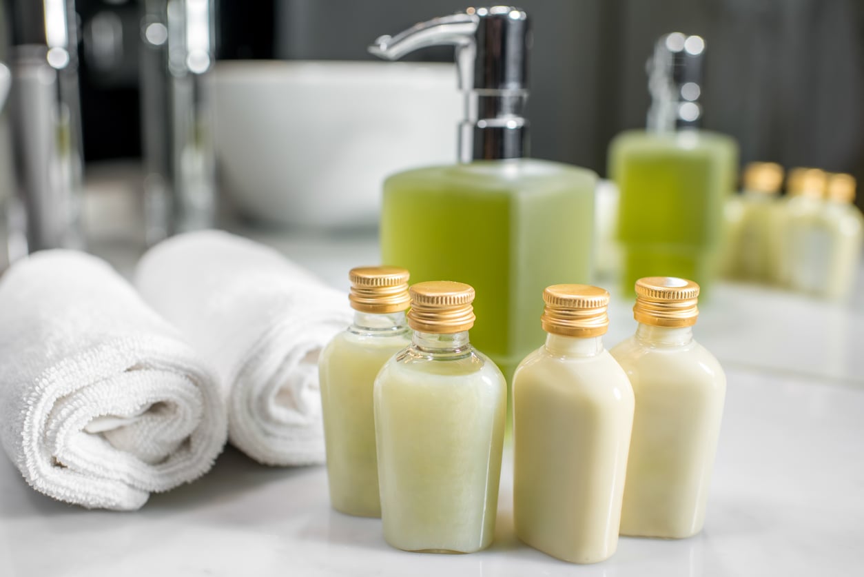 Four small bottles and a green soap dispenser are arranged on a bathroom counter, next to two neatly rolled white towels and a white bowl, creating a clean and spa-like setting—an inviting space designed to help you avoid common vacation mistakes by promoting relaxation and comfort throughout your stay.