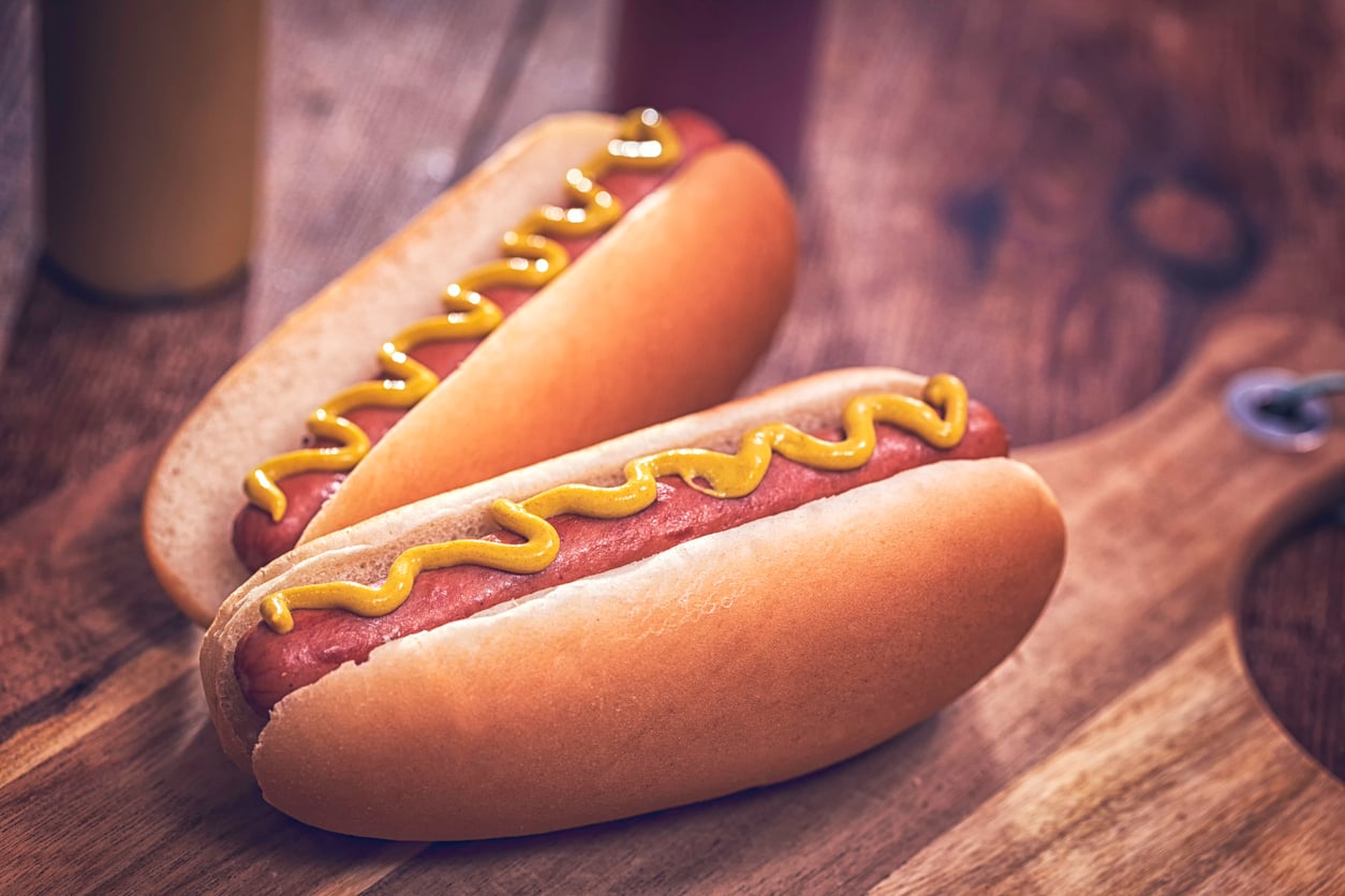 Two hot dogs in buns topped with mustard sit on a wooden surface, with a blurred background showing bottles, possibly of condiments—a classic example of foods people learn to appreciate later in life.