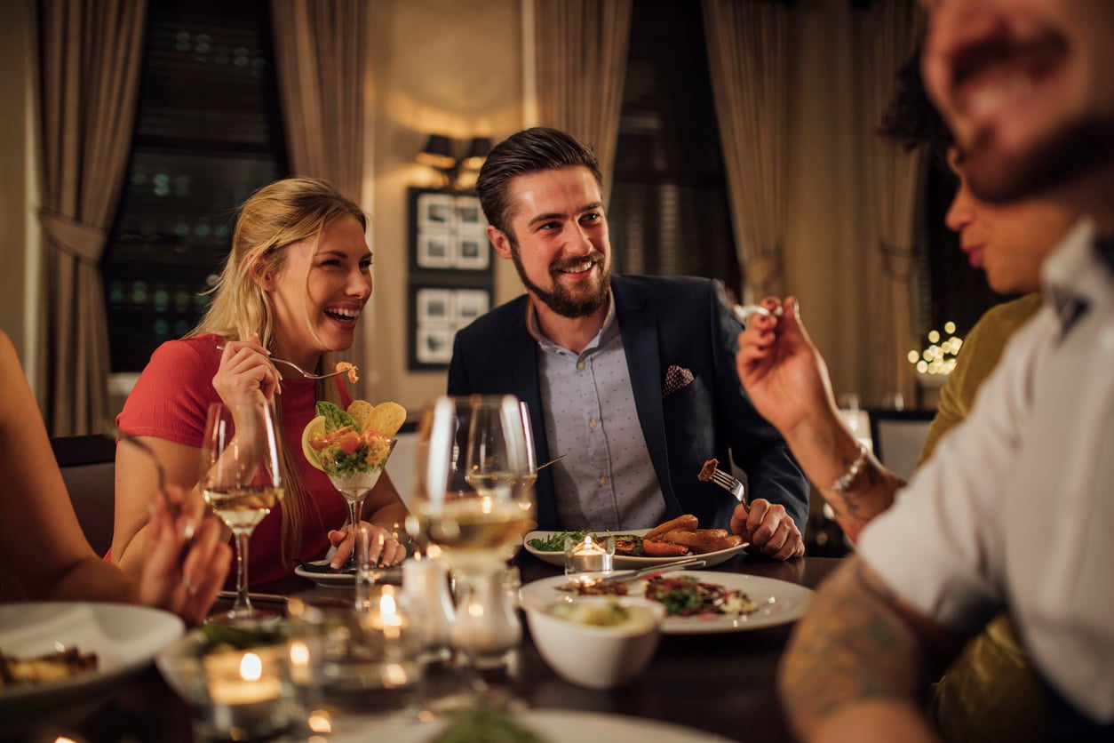 Four friends laugh and enjoy dinner together at a restaurant, surrounded by plates of food, glasses of wine, and candlelight. The atmosphere is warm and inviting.