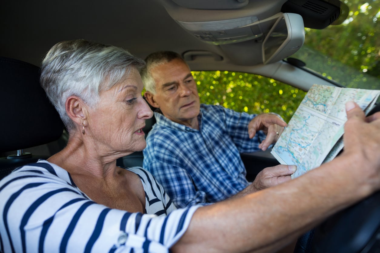 An older woman and man sit in a car, looking at a map together. The woman holds the map while the man points at it, appearing to discuss directions. Sunlight and greenery are visible through the window.