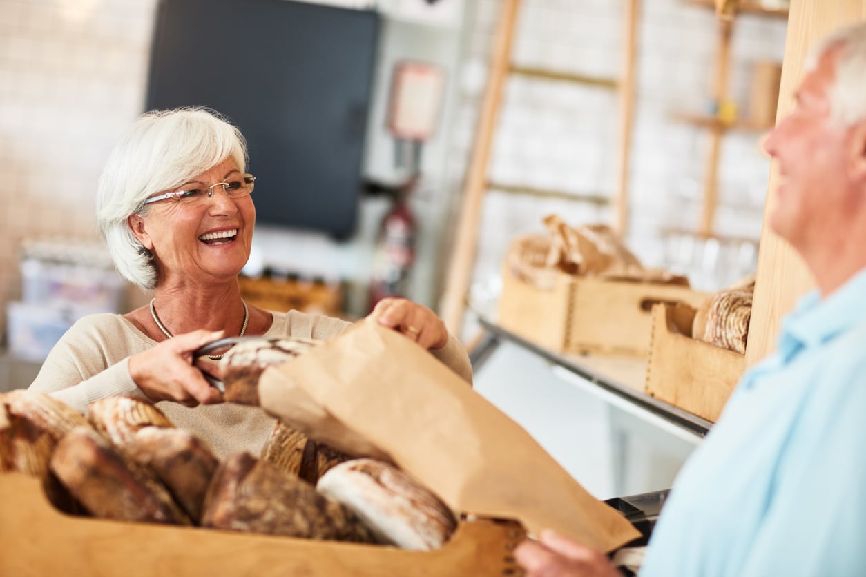 An older woman smiles as she places a loaf of bread into a paper bag for a customer in a bakery filled with various bread loaves.