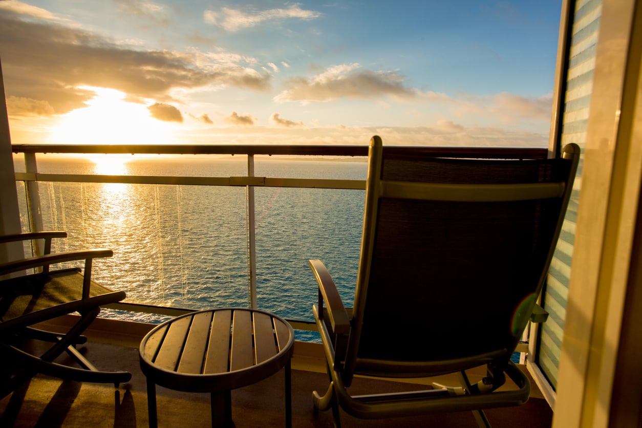 A balcony with two chairs and a small table overlooks a calm ocean at sunset, with warm sunlight reflecting on the water and clouds in the sky.