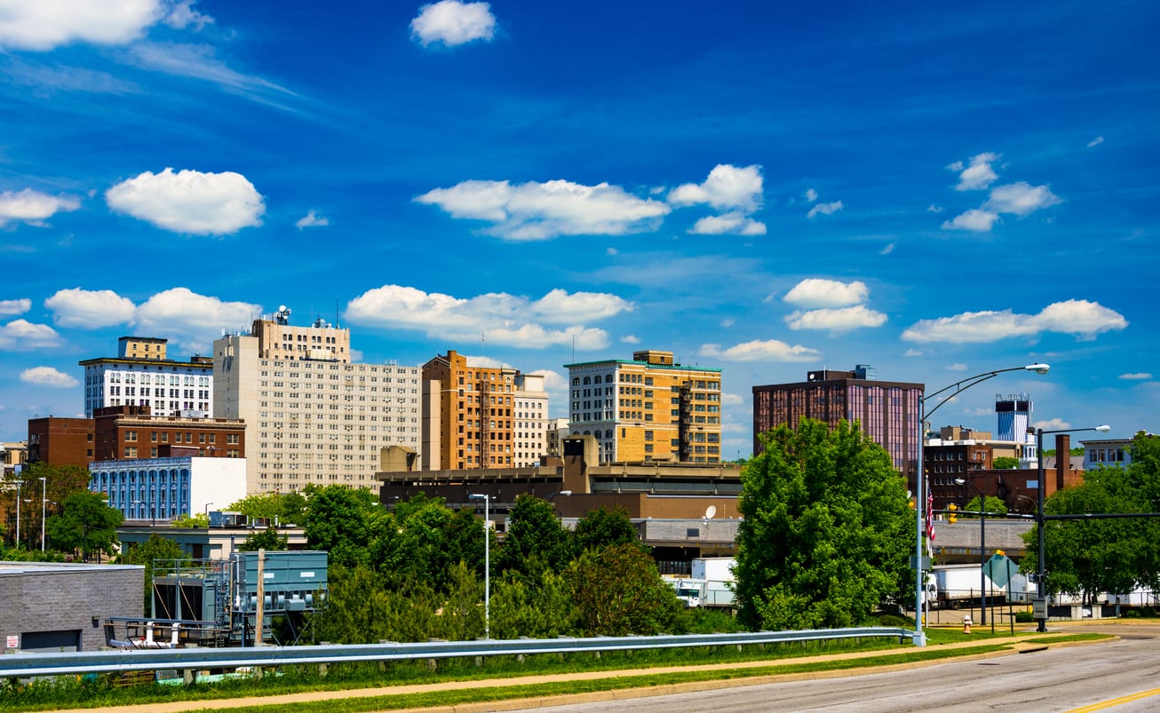 A city skyline with mid-rise buildings under a blue sky with scattered clouds, trees and greenery in the foreground, and a road with a metal guardrail at the bottom.