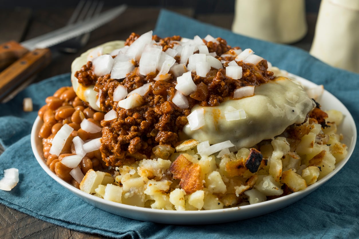 A plate of chopped potatoes, baked beans, and a cheeseburger patty topped with chili and raw chopped onions—one of our restaurant specialties—is served on a blue napkin with utensils in the background.