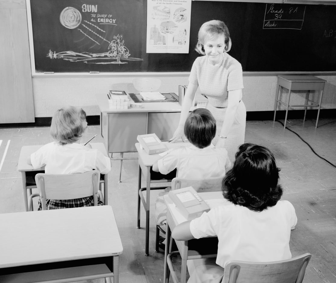 A teacher stands at a desk in front of three children seated at desks in a classroom. The chalkboard behind her has a drawing labeled “SUN The Source of All Energy.”