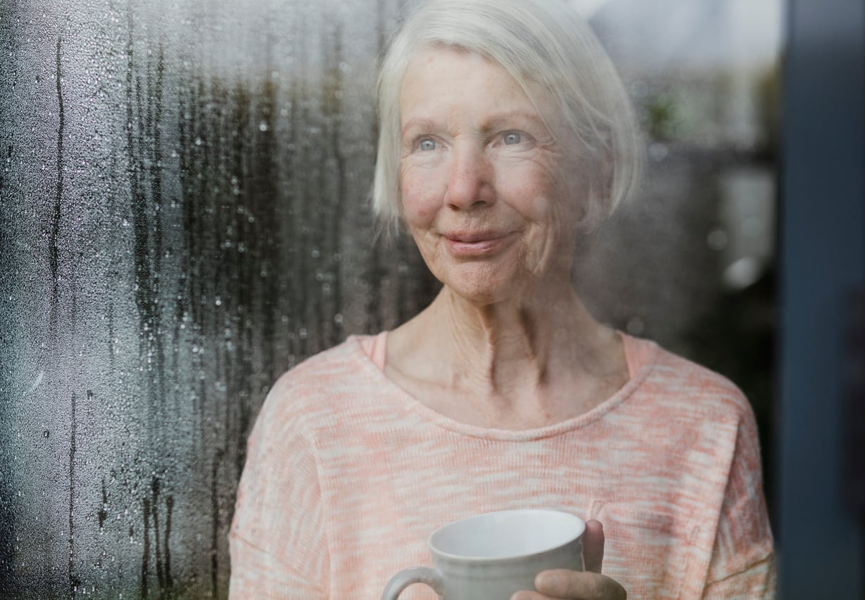 An older woman with short gray hair holds a mug and looks out a rain-streaked window, appearing thoughtful. She wears a light pink sweater and stands indoors, with soft daylight illuminating her face.