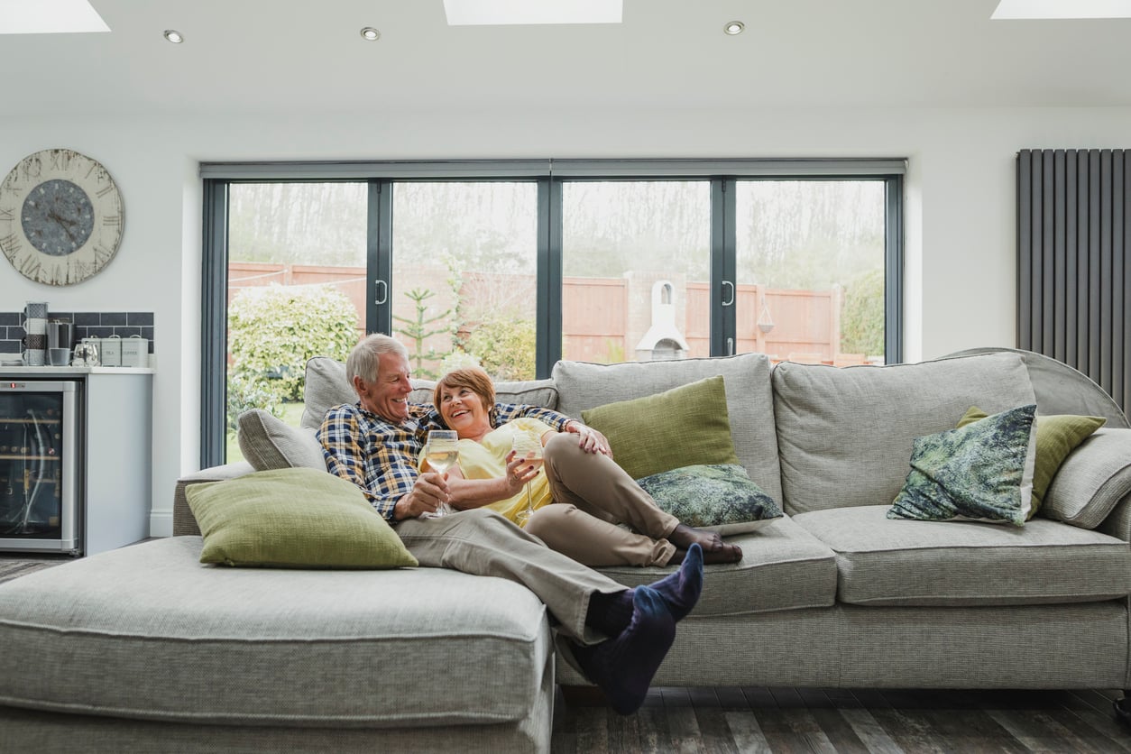 An older couple sits on a large grey sofa, smiling and embracing each other in a bright living room with large windows, green cushions, and a wall clock in the background.