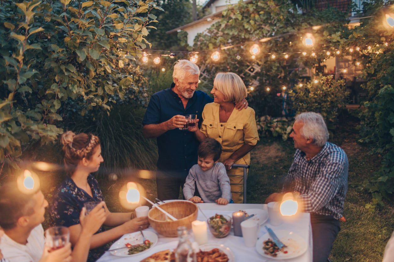 Six people, including a young child, enjoy a meal together outside at a garden table, surrounded by greenery and string lights, smiling and talking in a warm, festive atmosphere.