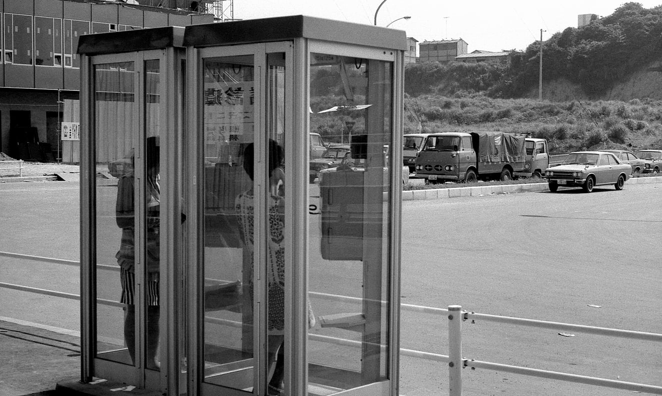 Two people stand inside a glass phone booth on a street, with parked vehicles and buildings visible in the background on a sunny day. Captured in black and white, the image evokes the atmosphere of travel in the 60s and 70s, when phone booths were essential for staying connected while exploring new places.