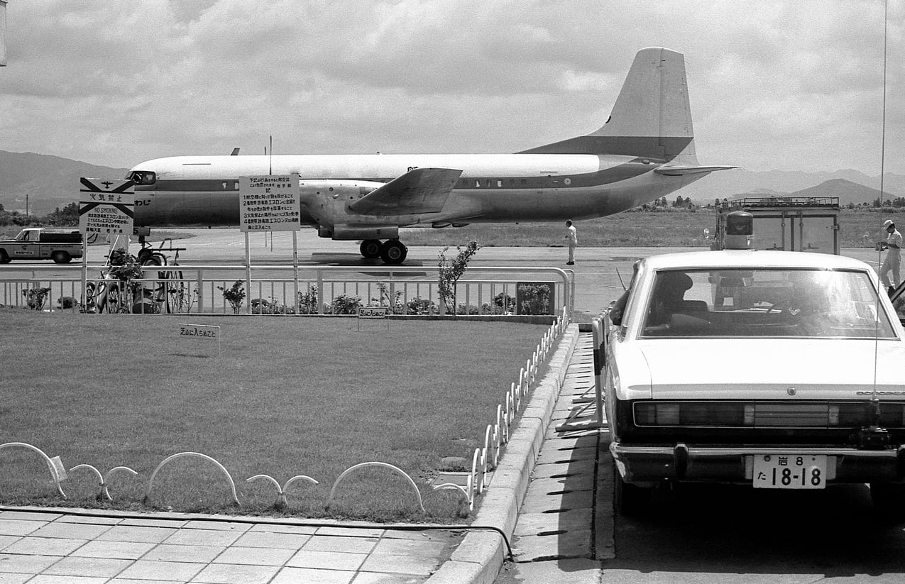 A large airplane on a runway is seen from behind a fence. In the foreground, a white car is parked next to grass and a sidewalk. People stand near the plane, and there are signs by the fence.