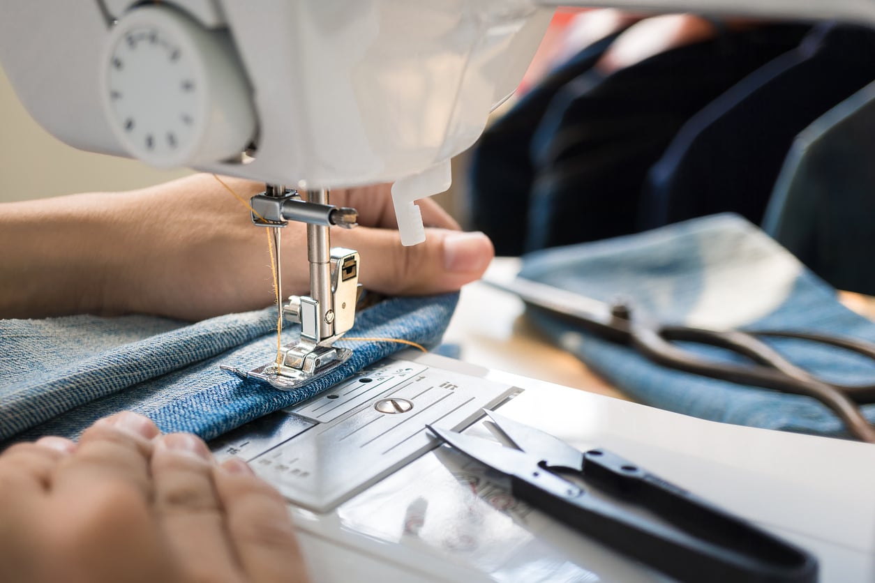A close-up of hands guiding denim fabric through a sewing machine, with small scissors and blue jeans in the background.