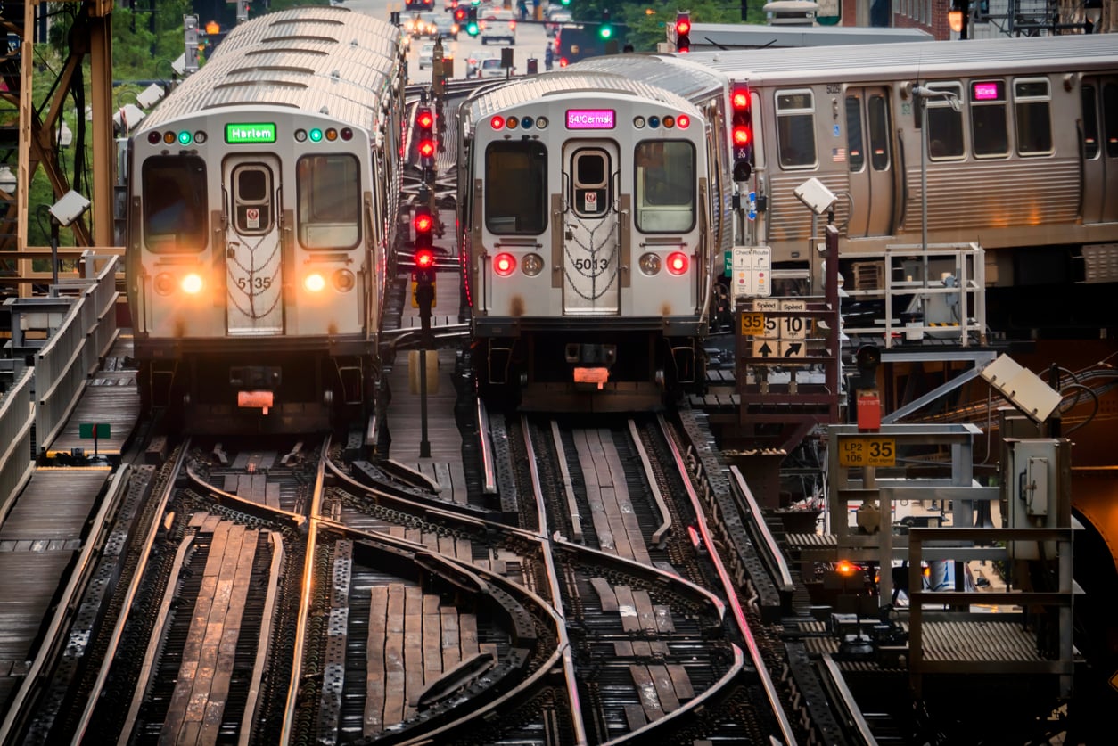 Two Chicago L trains approach each other on elevated tracks, surrounded by signals, lights, and city buildings in the background. Multiple tracks intersect, showing a busy urban transit scene.