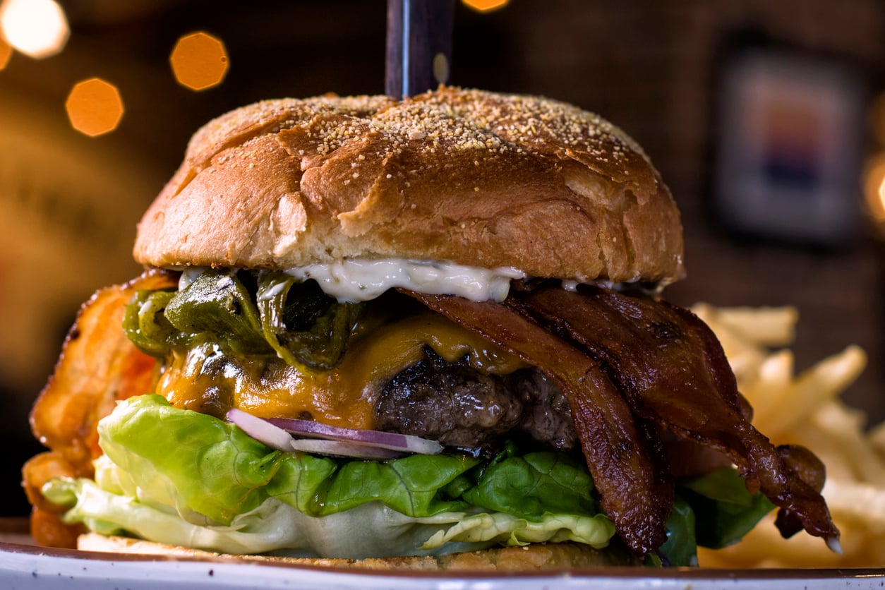 Close-up of a cheeseburger with bacon, lettuce, onions, cheese, pickles, and mayonnaise in a bun, served with fries in the background. A knife is sticking through the center of the burger.
