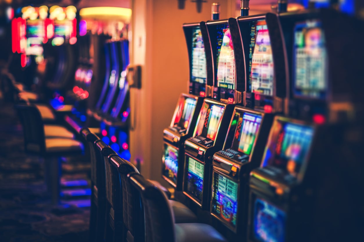 Rows of brightly lit slot machines with colorful screens line a casino floor. Empty chairs are positioned in front of the machines, and the atmosphere is dimly lit with neon and ambient lighting.