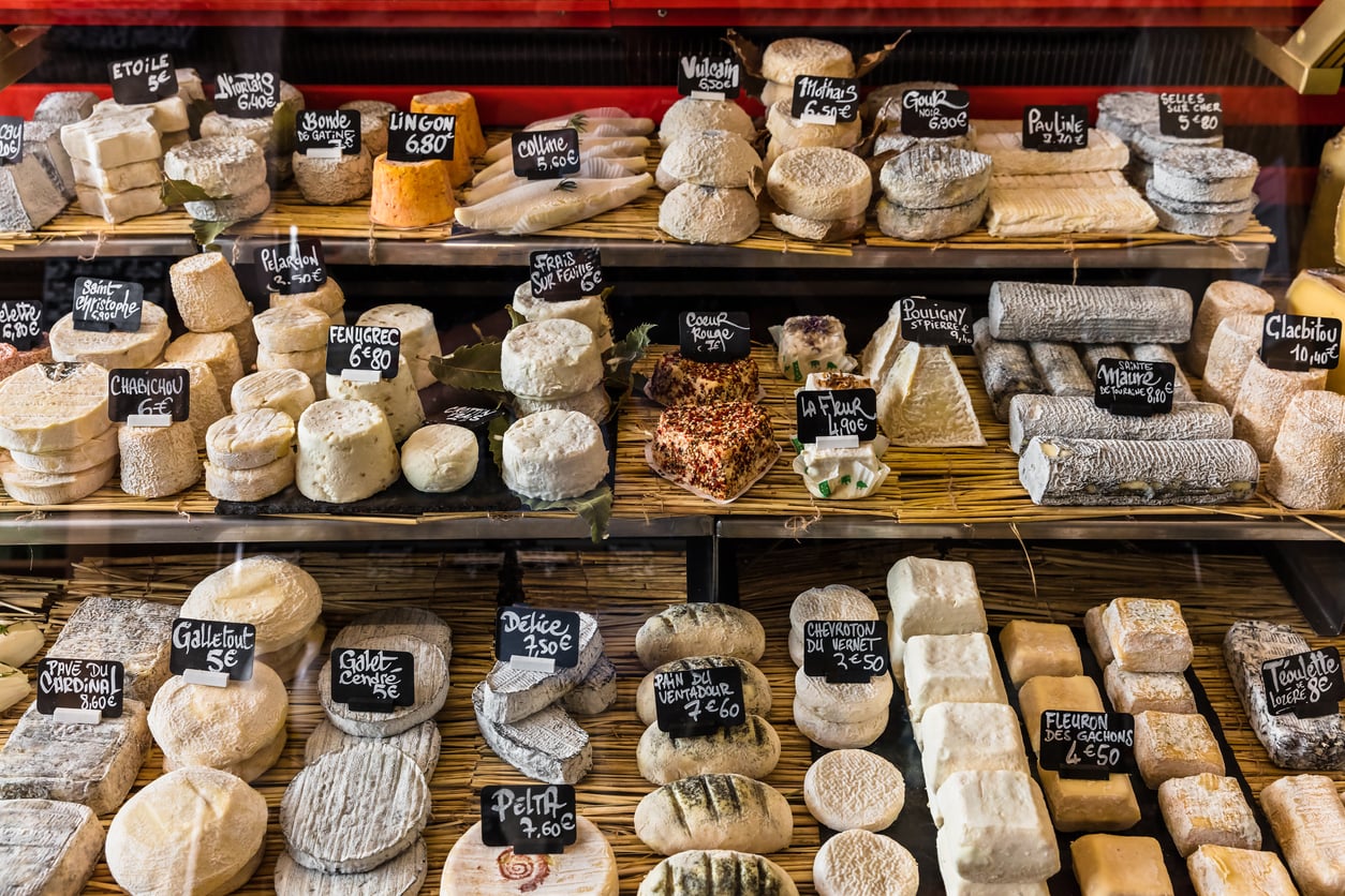 A cheese shop display with various shapes and sizes of cheeses, each labeled with small black signs showing names and prices, arranged on straw mats inside a glass case.