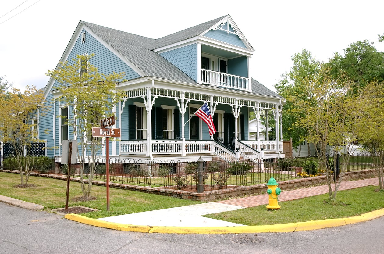 A blue Victorian-style house with a white porch, American flag, and a picket fence on a tree-lined street. A yellow fire hydrant and street signs are visible at the corner.