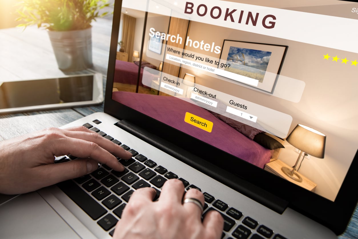 Person using a laptop with a hotel booking website on screen, showing fields to search hotels, check-in and check-out dates, number of guests, and photos of a hotel room.