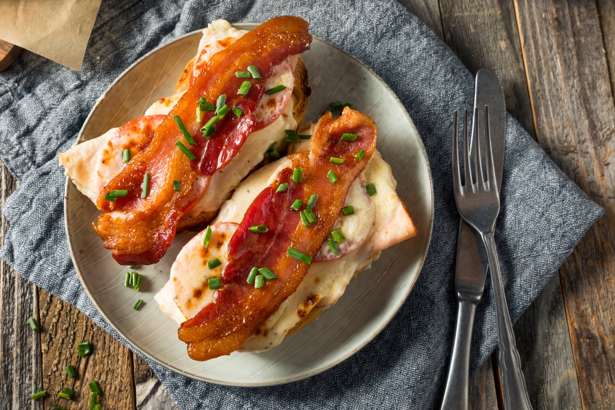 An open-faced sandwich topped with melted cheese, bacon, and chopped chives sits on a plate next to a fork and knife on a gray napkin and rustic wooden table.