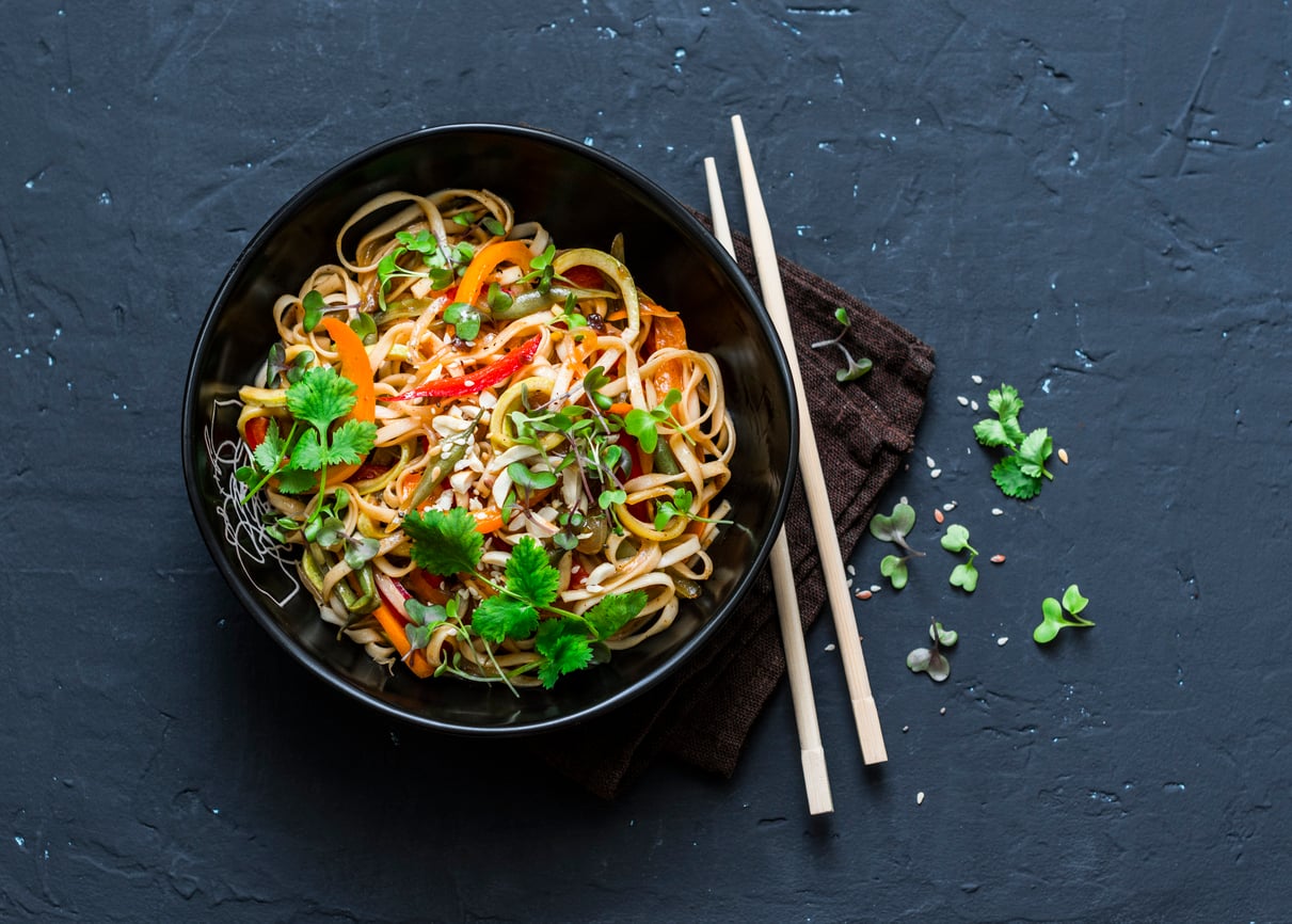 A black bowl of noodle salad with vegetables and cilantro, placed on a brown napkin, with a pair of chopsticks and some scattered greens on a dark textured surface—an inviting option among foods to order for a fresh and vibrant meal.