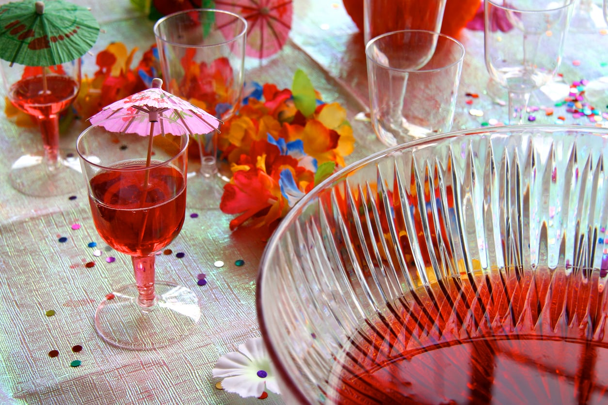 A punch bowl and glasses with red punch, colorful drink umbrellas, leis, and confetti on a festive table.