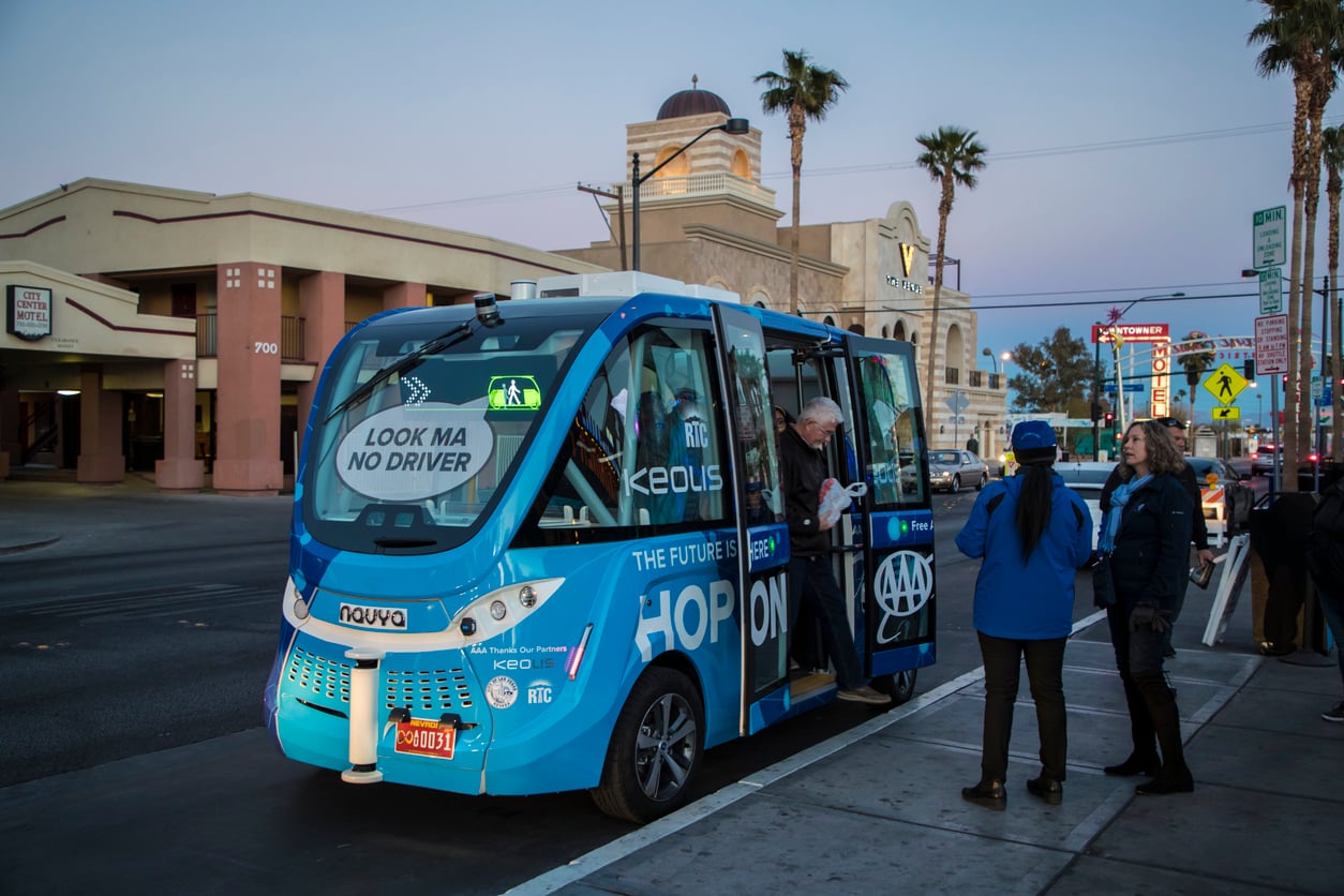 A blue self-driving shuttle with “LOOK MA NO DRIVER” on the windshield is parked on a street. Several people stand nearby, some boarding, with palm trees and buildings in the background.