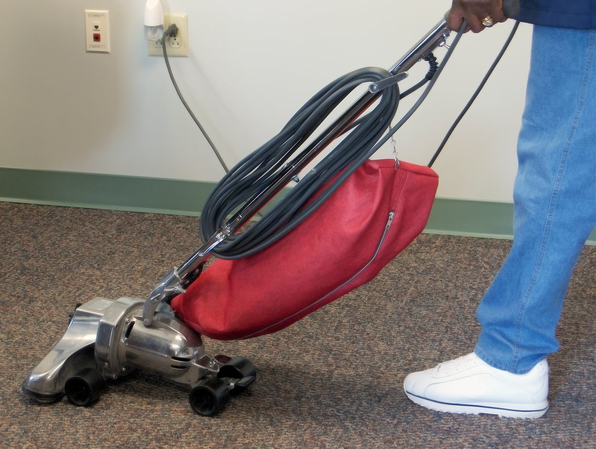A person in white sneakers and blue pants pushes a household items vacuum cleaner with a red dust bag on a carpeted floor near a white wall and electrical outlets.