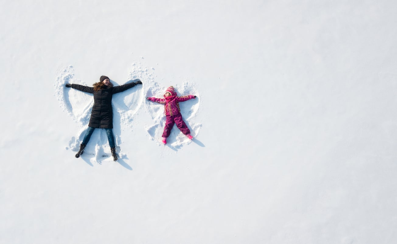An adult and a child in winter clothes lie on their backs in the snow, making snow angels. The scene is bright, with untouched white snow surrounding them.