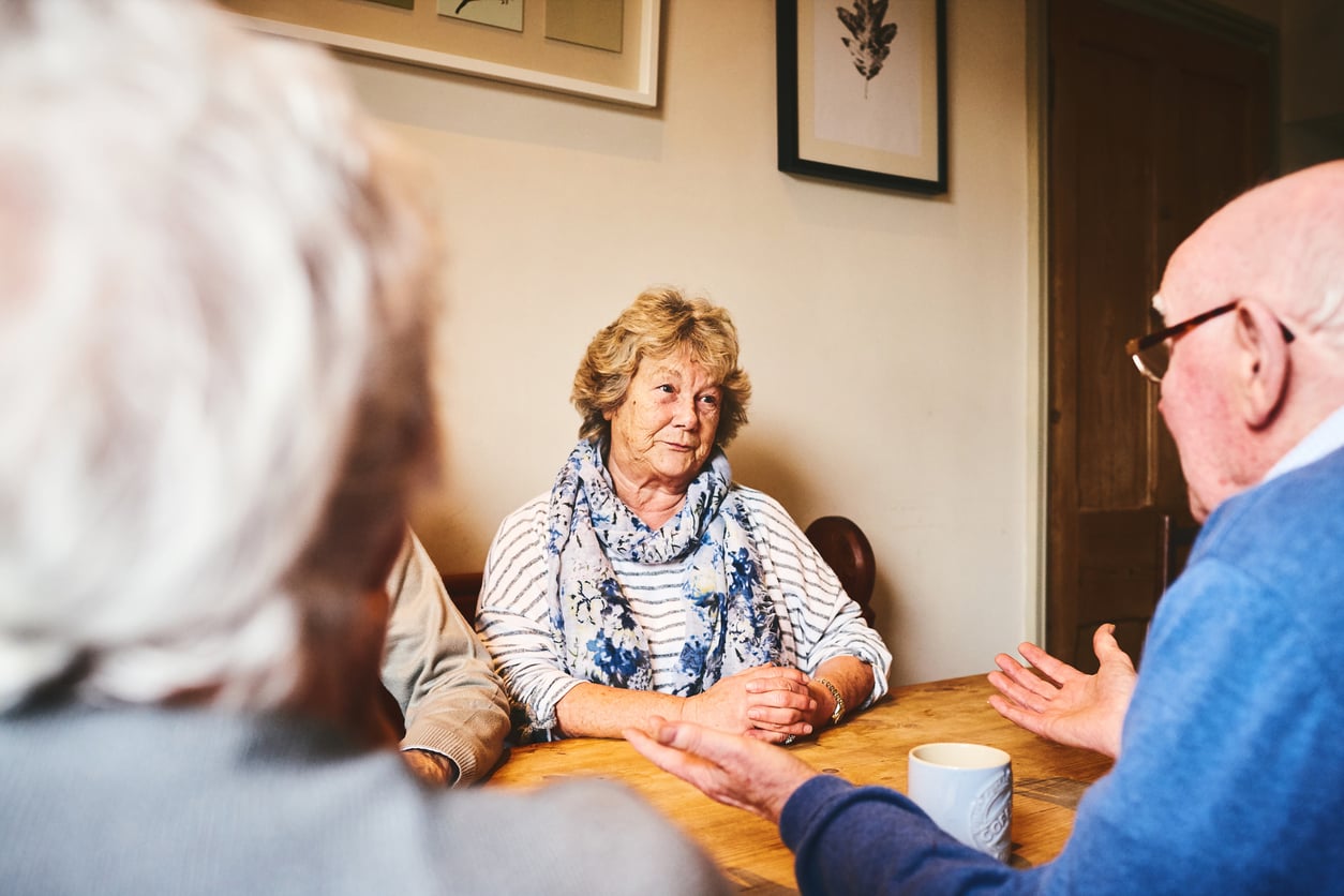 Three older adults sit around a wooden table, engaged in conversation. A woman in the center smiles, listening to a man with glasses who gestures with his hands. Paintings hang on the wall behind them.