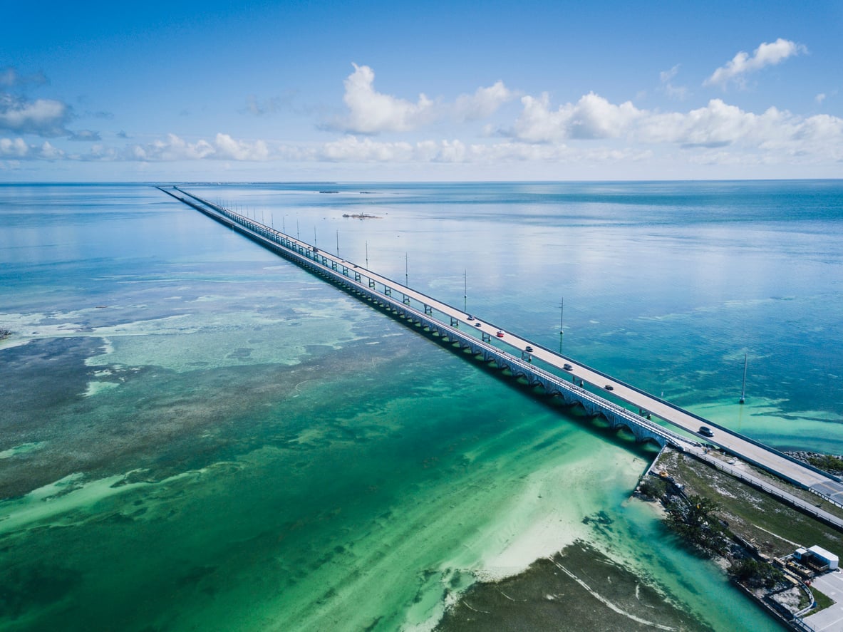 A long bridge stretches across turquoise water under a blue sky with scattered clouds, cars traveling along it, connecting small islands in the distance.