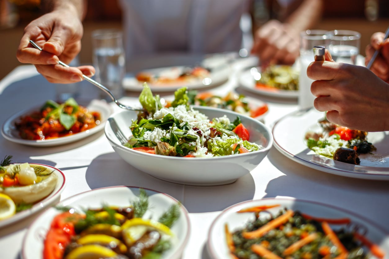 People sharing a meal outdoors with plates of colorful salads, grilled vegetables, and a large bowl of leafy green salad in the center of the table. Hands are seen serving food.