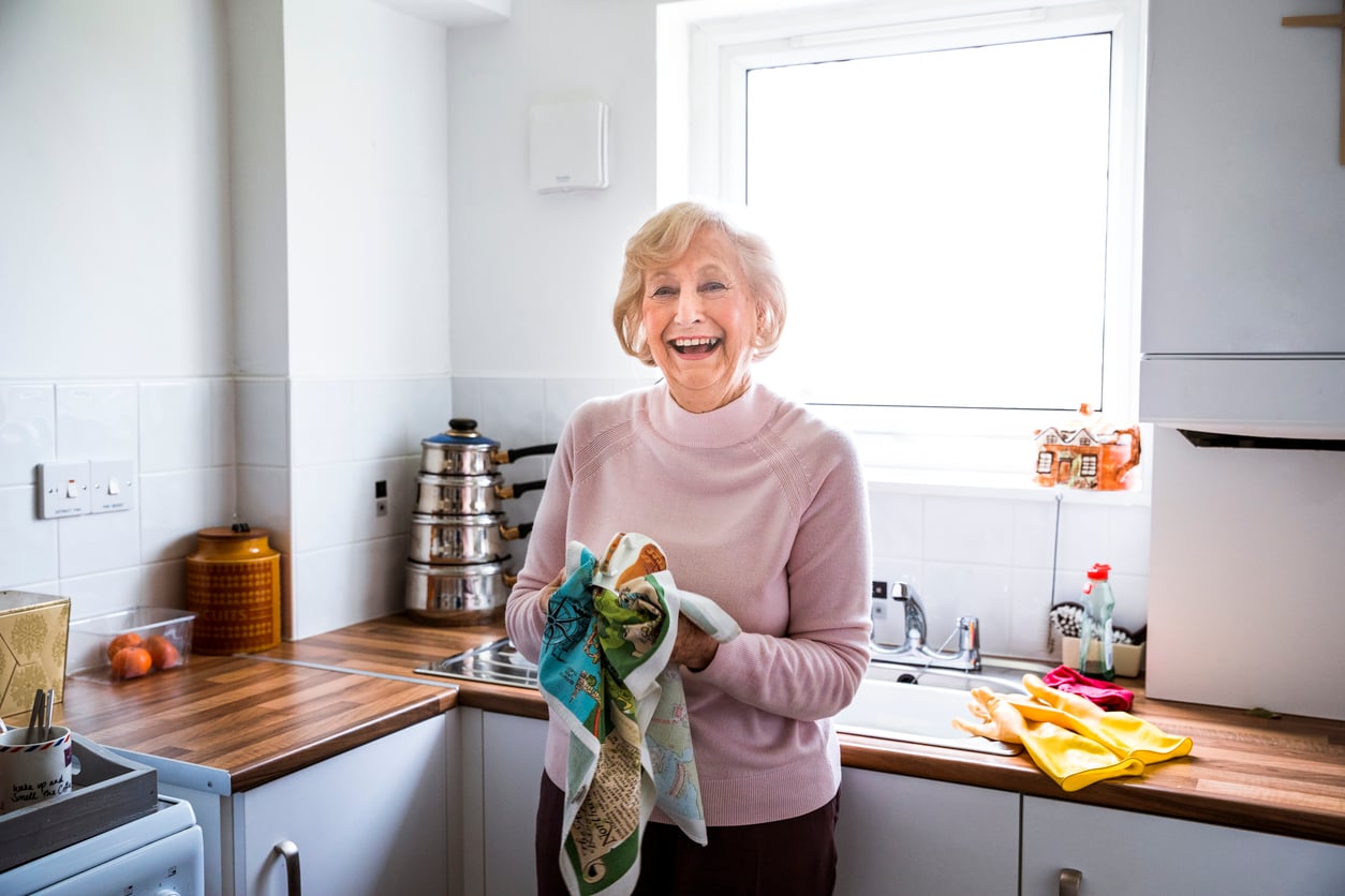 An older woman with short blonde hair smiles while drying dishes with a towel in a bright, modern kitchen.