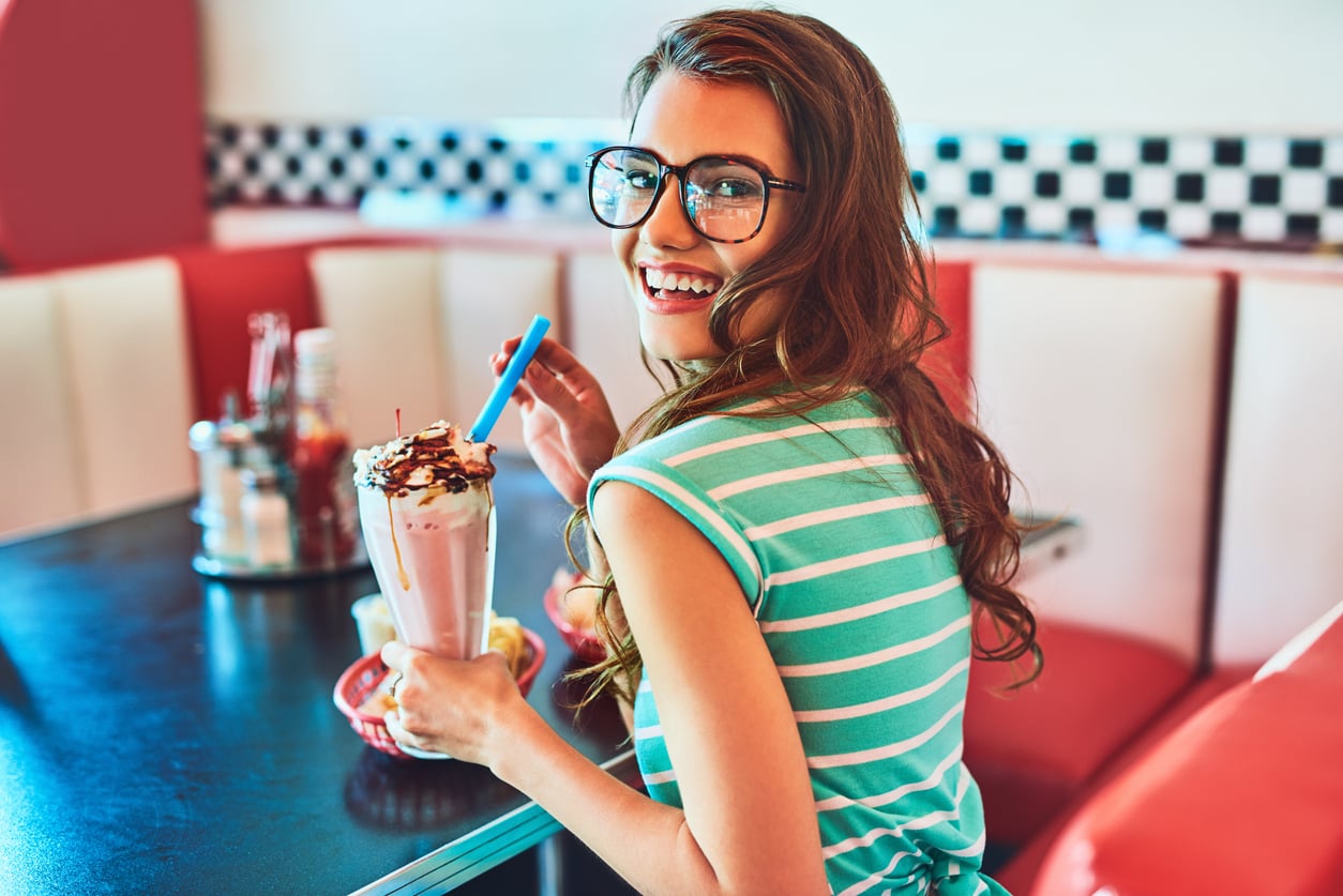 A smiling woman in glasses and a striped shirt sits in a retro diner, holding a milkshake topped with whipped cream and chocolate, looking back at the camera.
