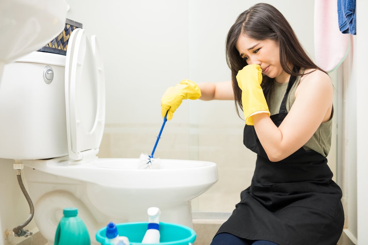 A woman wearing yellow gloves and an apron cleans a toilet with a brush while holding her nose, looking uncomfortable. Cleaning supplies are in a bucket on the floor nearby.
