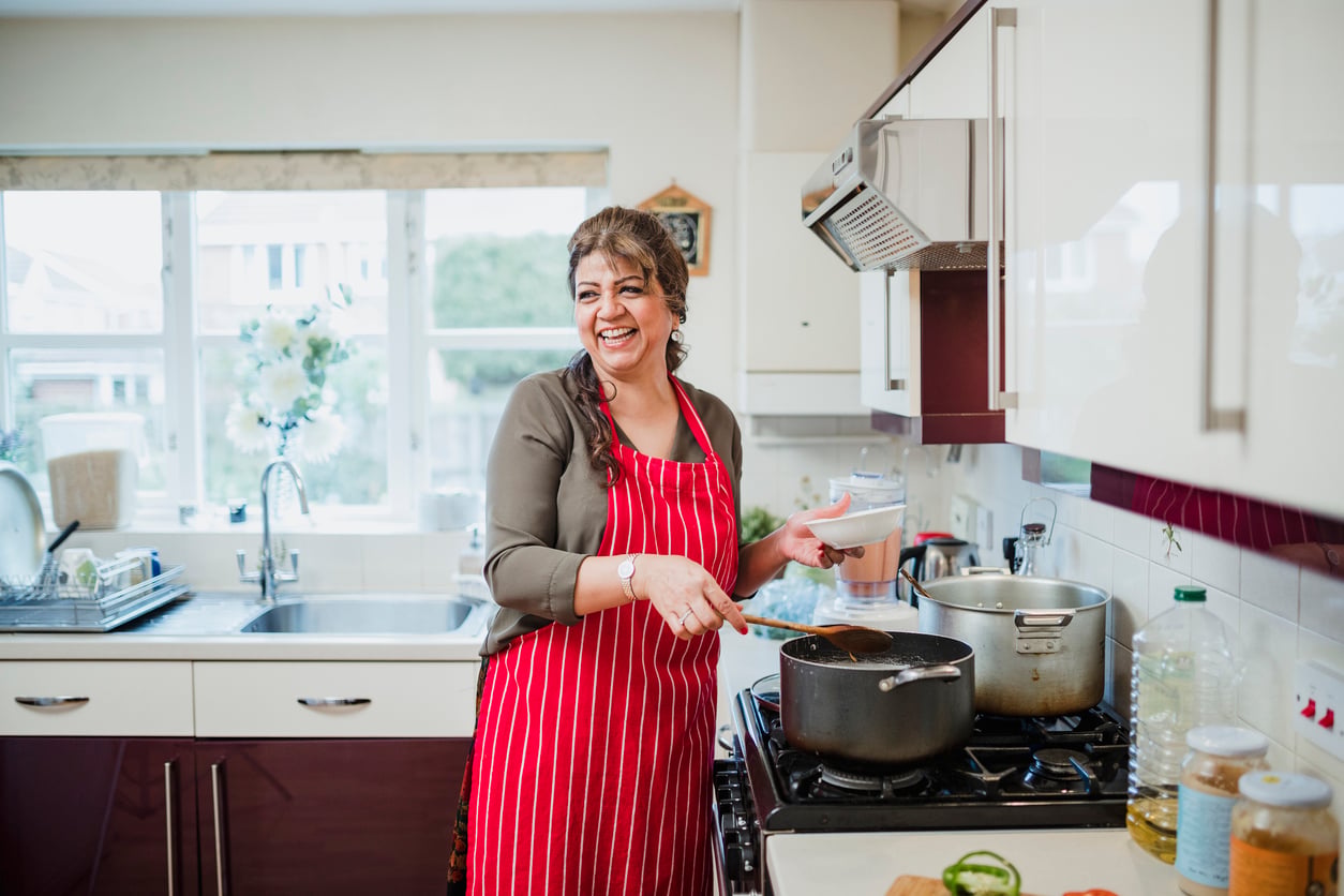 A woman in a red striped apron smiles while cooking at a stove in a bright kitchen, holding a spoon and a small bowl, with pots and vegetables nearby.