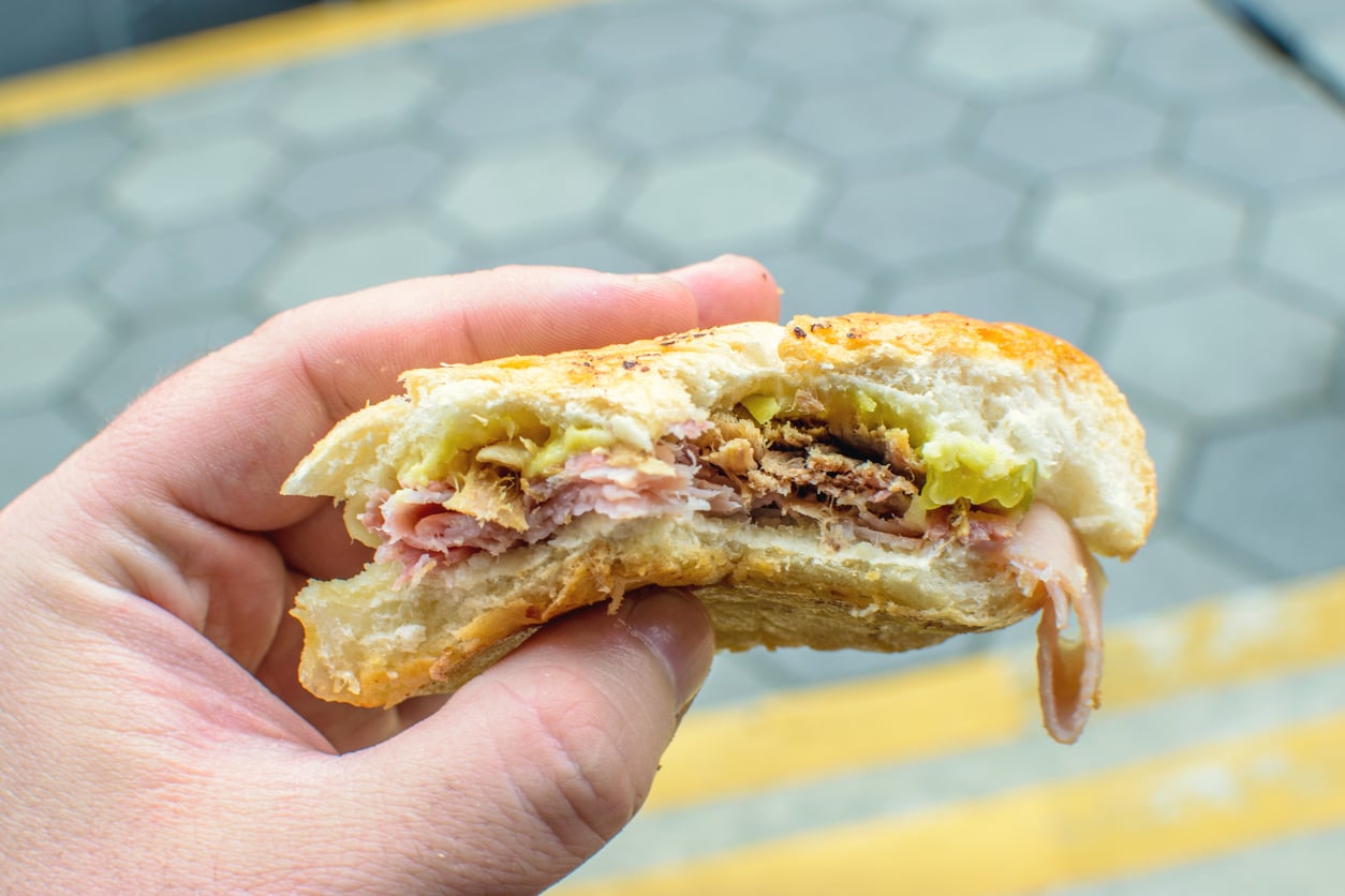 A hand holding a half-eaten sandwich filled with meats, lettuce, and possibly pickles, with a blurred sidewalk and yellow lines in the background.