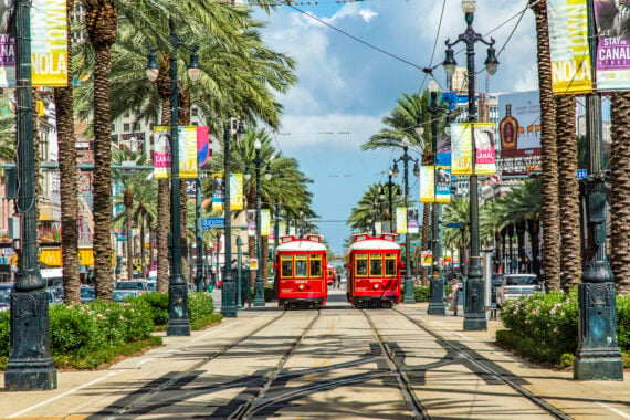 Two red streetcars travel down palm tree-lined tracks in New Orleans, with colorful &ldquo;NOLA&rdquo; banners and city buildings visible under a sunny sky.