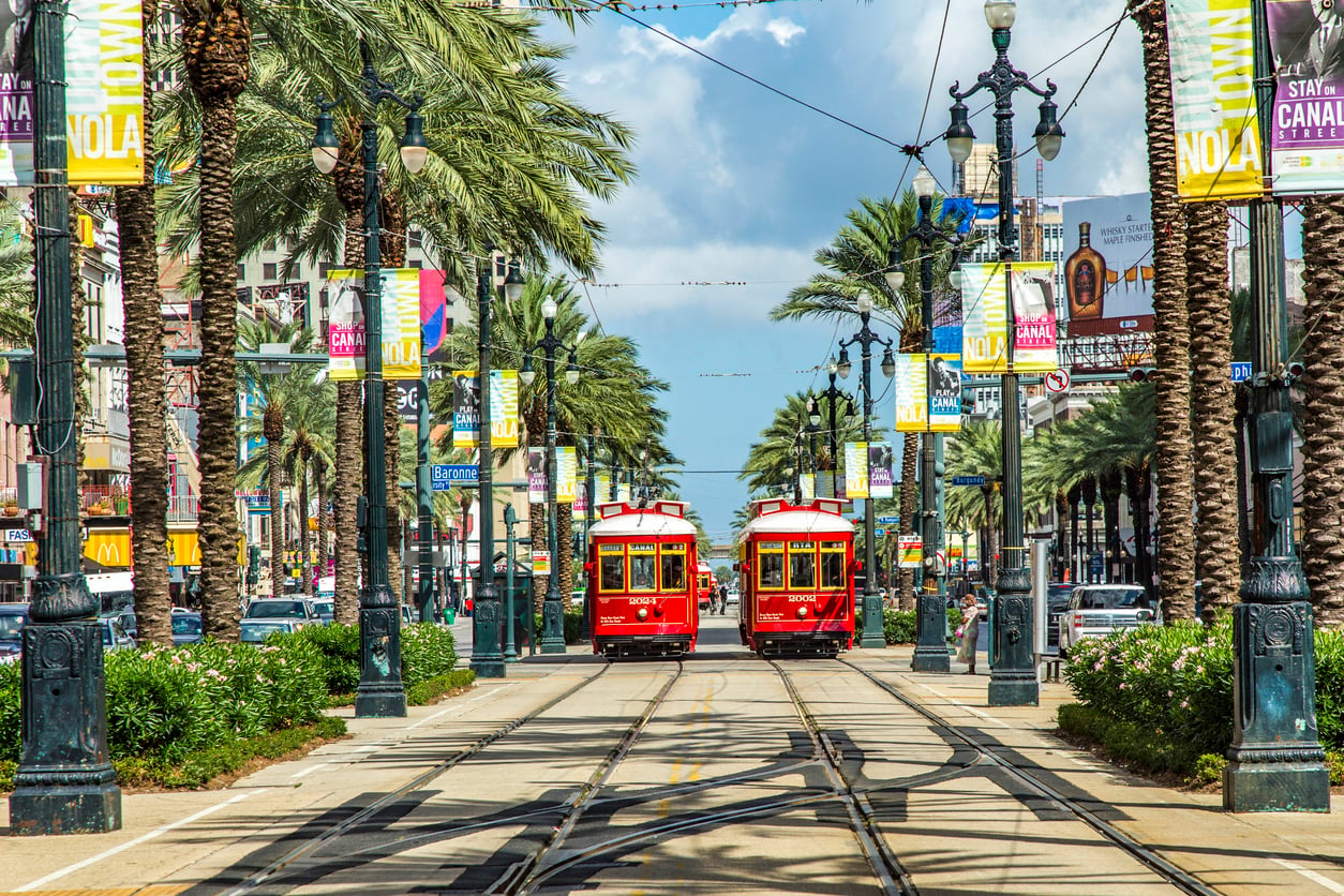 Two red streetcars travel down palm tree-lined tracks in New Orleans, with colorful “NOLA” banners and city buildings visible under a sunny sky.