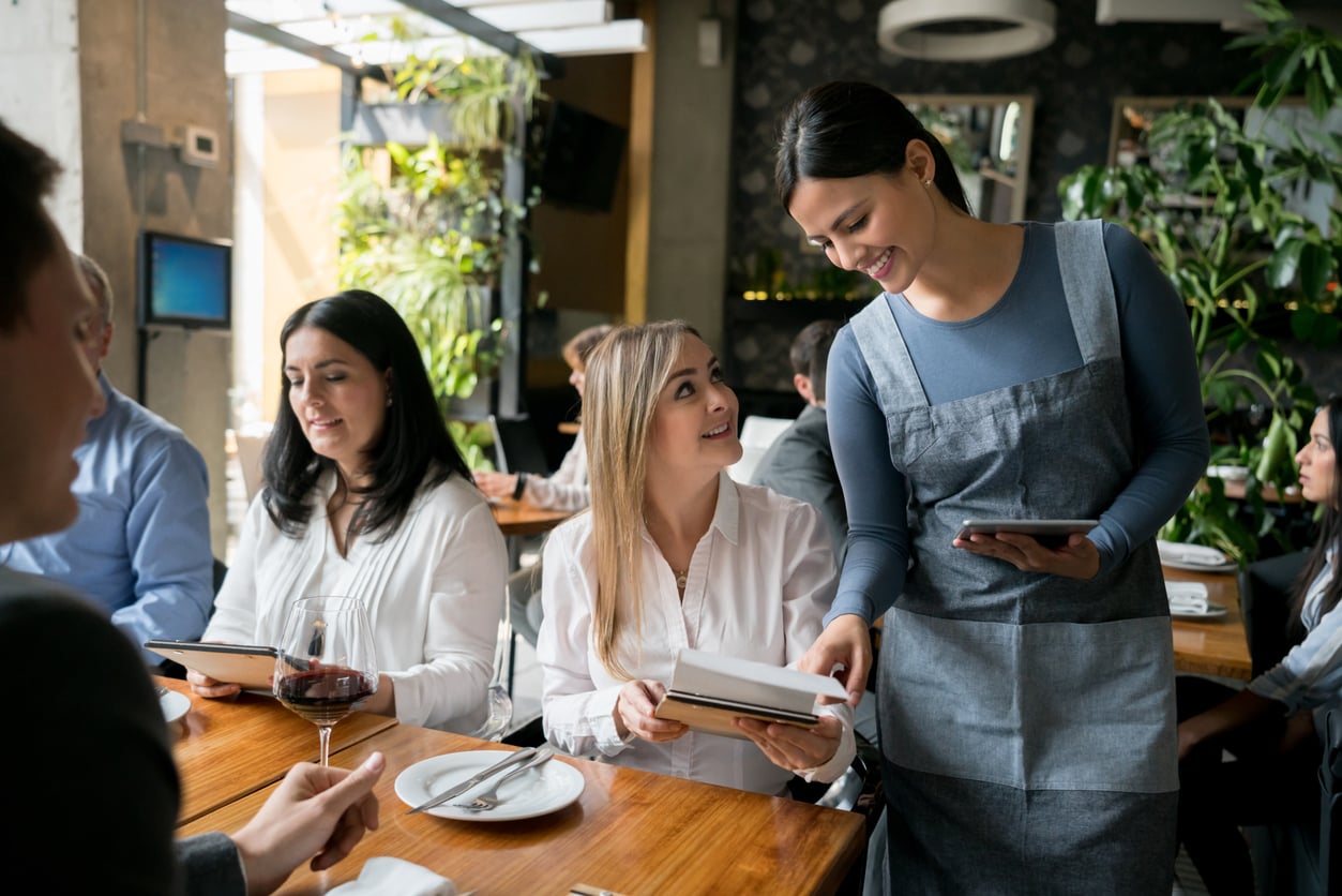 A smiling waitress takes an order from a group of people seated at a restaurant table, with one woman holding a menu and looking up at the waitress.