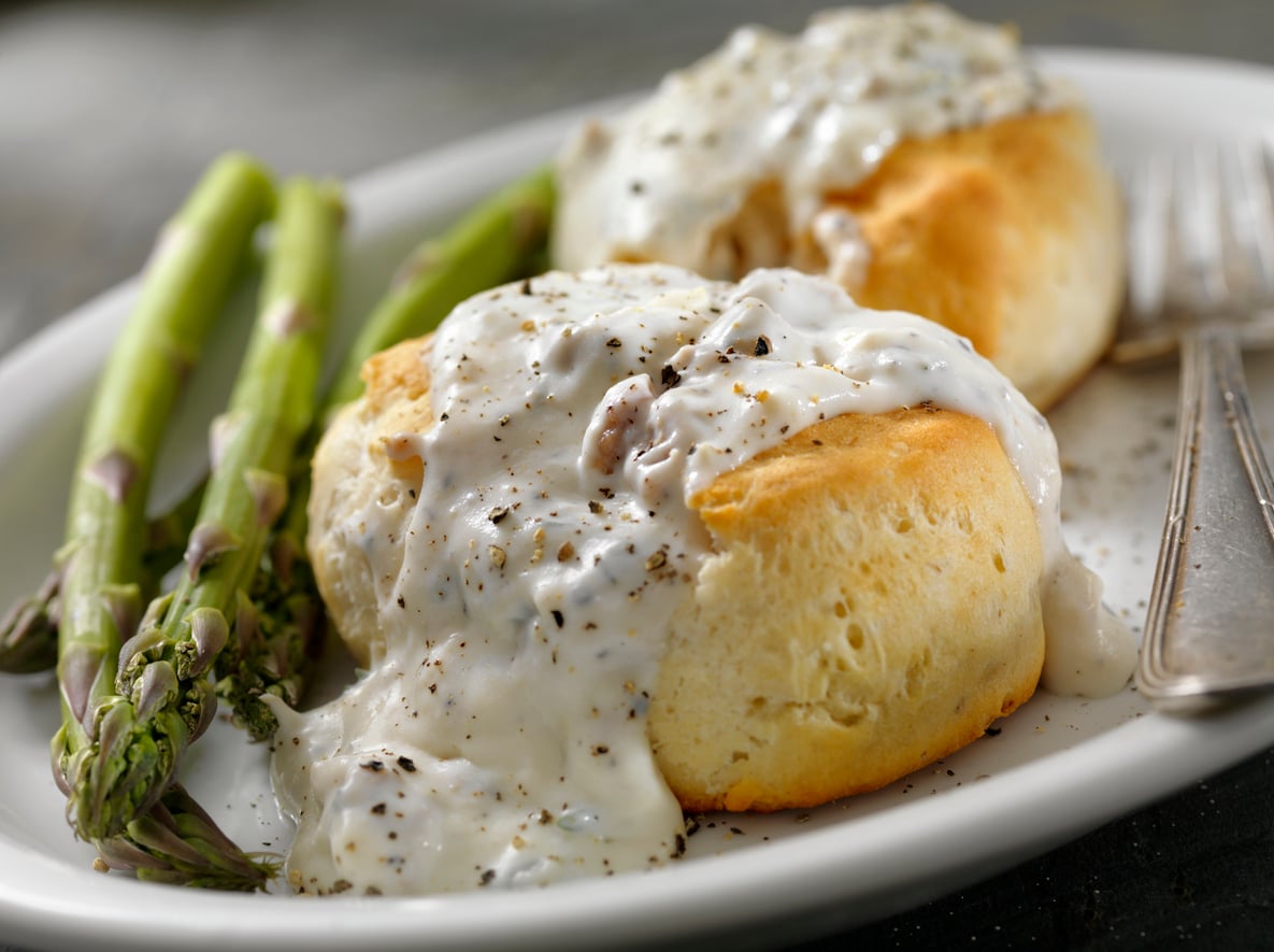 Two biscuits topped with white sausage gravy and black pepper on a white plate, served with asparagus spears and a fork.