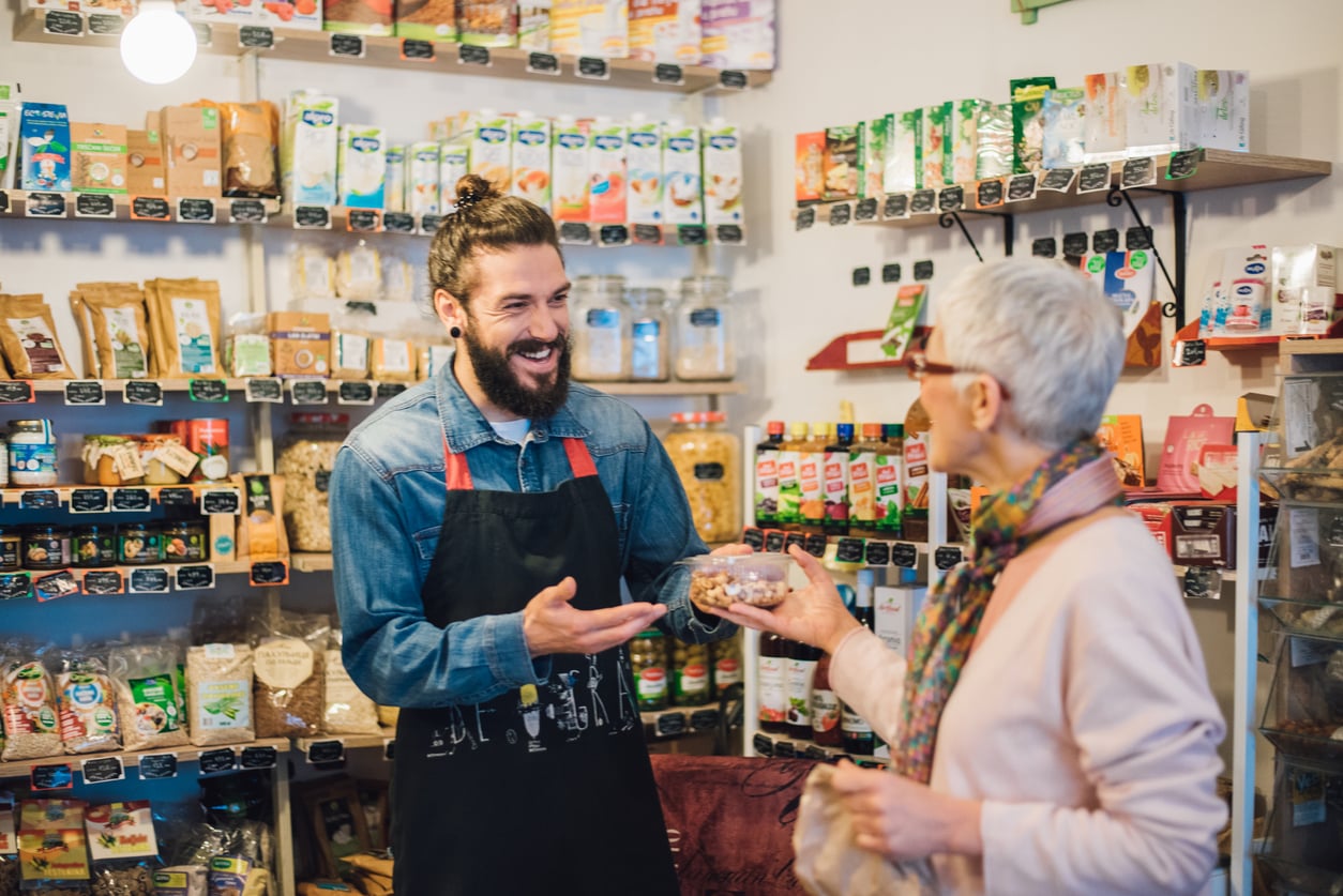 A smiling shopkeeper hands a food container to a customer in a small grocery store filled with various packaged goods on shelves.