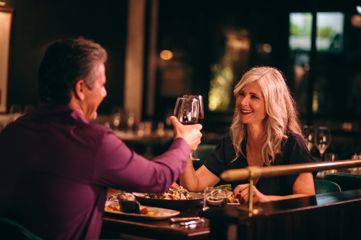 A man and woman sit across from each other at a restaurant table, smiling and raising glasses of red wine for a toast, with plates of food in front of them in a warmly lit setting—a special treat as dining out has become one of the things families spend less on.