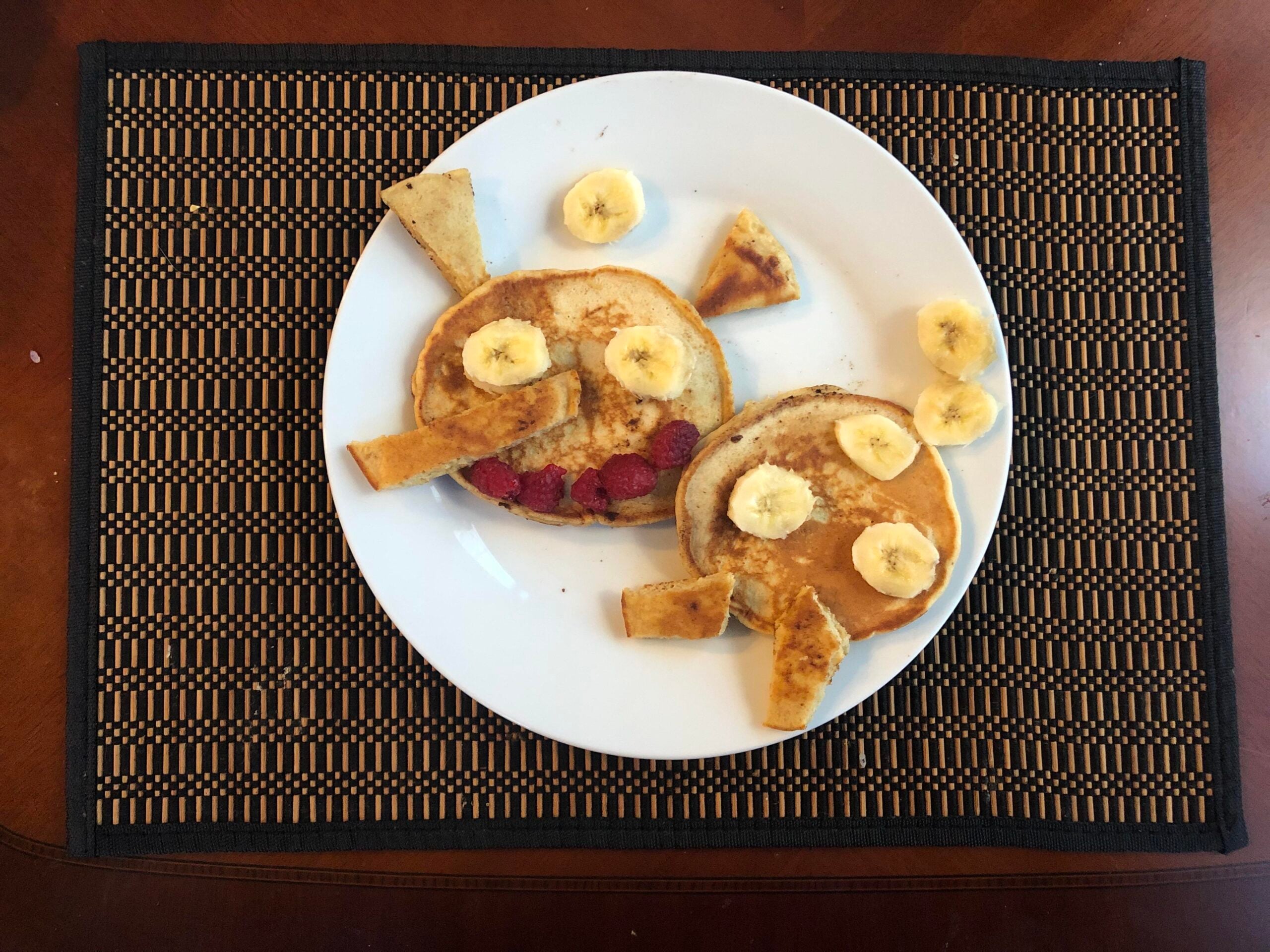 Two pancakes on a white plate are decorated to look like fish using banana slices, raspberries, and toast pieces. The plate sits on a black and tan woven placemat on a wooden table.